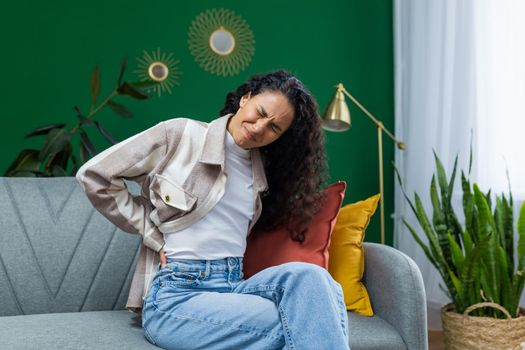 Young beautiful hispanic woman at home having severe back pain, housewife sitting on sofa in living room and massaging back side in room with green wall.