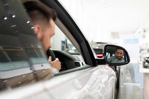 a man at a car dealership driving a car looks in the rearview mirror.