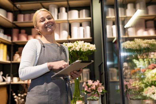 Satisfied woman self-employed owner of a flower shop with a tablet in her hands next to a refrigerator with fresh flowers.