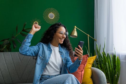 Happy and smiling hispanic woman at home is happy and smiling reading online message from smartphone, holding hand up victory gesture, celebrating triumph and success