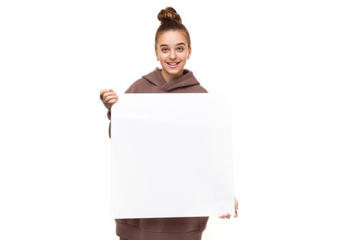 Photo of a beautiful positive smiling cute girl with dark hair and a bun in a brown hoodie with hood holding a large white square sheet with blank space isolated on white background with free space for text.