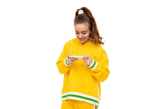 Pretty beautiful smiling cheerful cute girl with dark hair with a ponytail in a stylish yellow tracksuit with green stripes plays a mobile phone with a white case isolated on a white background with free space for text.