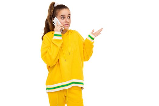 Photo of a beautiful shocked and surprised girl with dark hair with a ponytail in a stylish yellow tracksuit with green stripes talking on a mobile phone isolated on white background with free space for text.