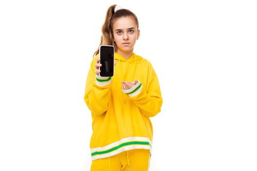 Photo of a beautiful girl with dark hair with a ponytail in a stylish yellow tracksuit with green stripes shows a mobile phone with a black blank display isolated on a white background with free space for text.