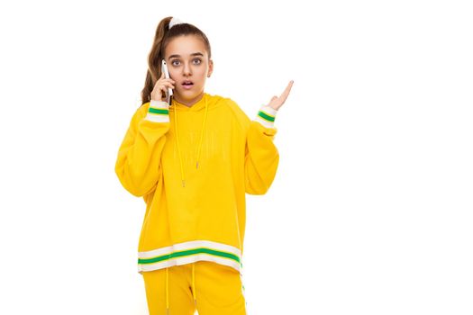 Photo of a beautiful shocked and surprised girl with dark hair with a ponytail in a stylish yellow tracksuit with green stripes talking on a mobile phone isolated on white background with free space for text.