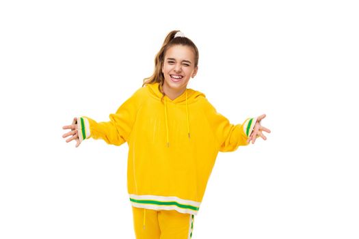 Photo of a beautiful charming smiling cute girl with dark hair with a ponytail in a stylish yellow tracksuit with green stripes shrugs her hands isolated on a white background with free space for text.