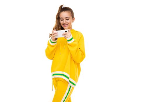 Photo of a beautiful smiling cheerful cute girl with dark hair with a ponytail in a stylish yellow tracksuit with green stripes plays a mobile phone with a white case isolated on a white background with free space for text.