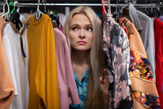 Girl worried because she hasn't found the dress she dreamed of. A blonde girl among hangers of clothes with a grim face.