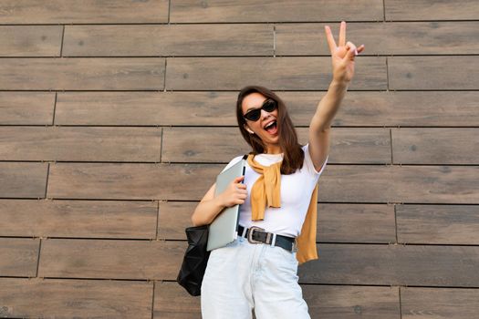 Photo of fascinating beautiful smiling charming young brunette girl wearing stylish clothes holding computer laptop and black sunglasses in white t-shirt and light blue jeans in the street near brown wall and showing peace gesture with two fingers.