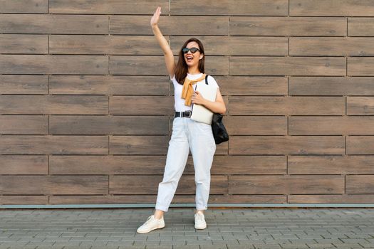 Photo of fascinating beautiful smiling charming young brunette girl wearing stylish clothes holding computer laptop and black sunglasses in white t-shirt and light blue jeans in the street near brown wall and handing up.