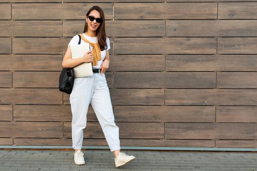 Photo of fascinating beautiful smiling charming young brunette girl wearing stylish clothes holding computer laptop and black sunglasses in white t-shirt and light blue jeans in the street near brown wall.