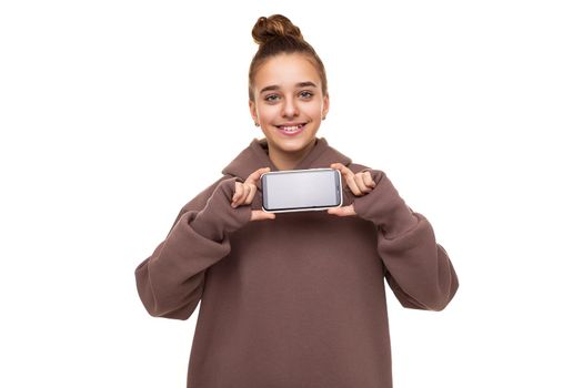 Photo of a beautiful cute girl with dark hair and a bun in a brown hoodie with a hood holding a mobile phone with a blank black screen isolated on a white background with free space for text.