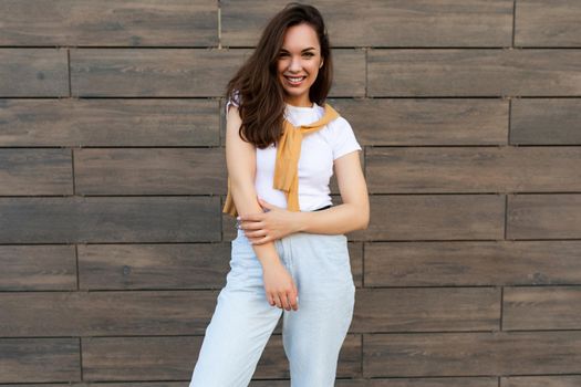 Photo of pretty attractive beautiful smiling happy positive charming young brunette young woman wearing stylish clothes: white t-shirt, yellow sweater and light blue jeans in the street near brown wall.