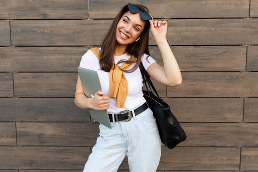 Photo of fascinating beautiful smiling happy positive charming young brunette girl wearing stylish clothes holding computer laptop and black sunglasses in white t-shirt and light blue jeans in the street near brown wall.