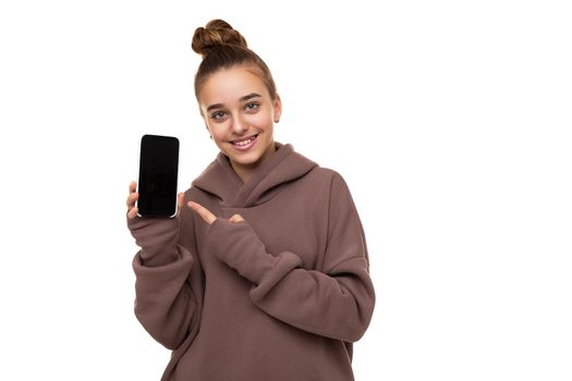 Photo of a beautiful cute girl with dark hair and a bun in a brown hoodie with a hood holding a mobile phone with a blank black screen isolated on a white background with free space for text.