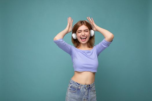 Charming happy smiling young blonde woman wearing blue crop top isolated on blue background with copy space for text wearing white wireless bluetooth headphones listening to good music dancing and having fun.