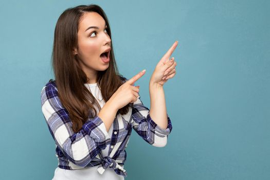 Young amazed, surprised, shocked beautiful brunette woman with sincere emotions wearing trendy check shirt standing isolated on blue background with empty space for text and showing to free space.