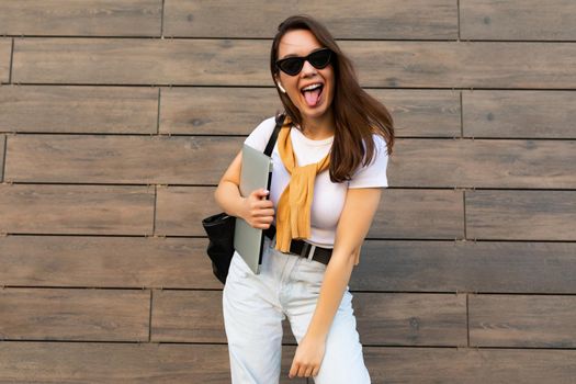 Photo of fascinating beautiful smiling charming young brunette girl wearing stylish clothes holding computer laptop and black sunglasses in white t-shirt and light blue jeans in the street near brown wall.
