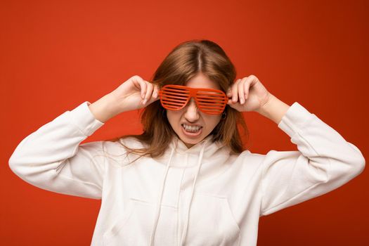 Photo shot of attractive positive happy young blonde woman isolated over colourful background with copy space for text wearing casual white hoodie and stylish glasses looking at camera and having fun.