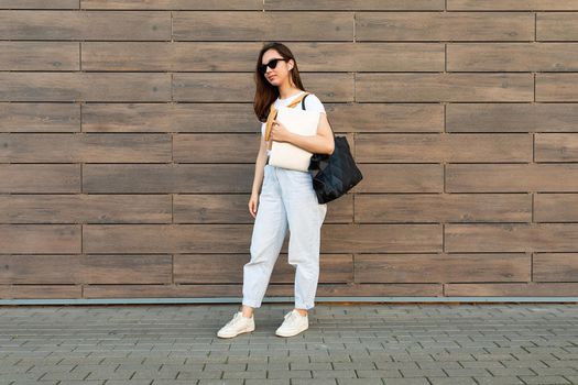 Photo of fascinating beautiful smiling charming young brunette girl wearing stylish clothes holding computer laptop and black sunglasses in white t-shirt and light blue jeans with black bag in the street near brown wall.
