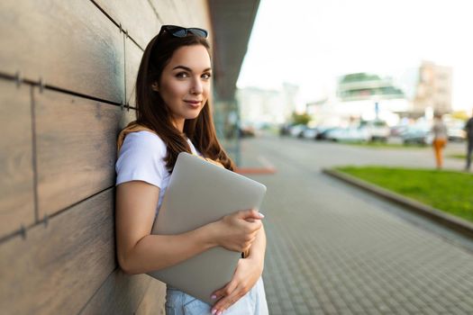 Photo of pretty attractive beautiful smiling positive charming young brunette young woman holding gray computer laptop wearing stylish clothes in white t-shirt and light blue jeans in the street in the town near brown wall.