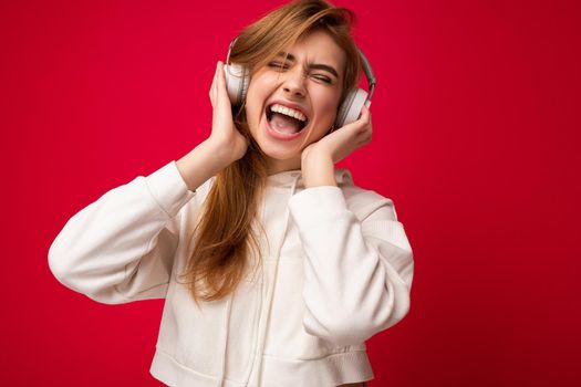 Photo of beautiful happy smiling young blonde woman wearing white hoodie isolated over colourful background wall with copy space for text wearing white wireless bluetooth earphones listening to cool music and having fun.