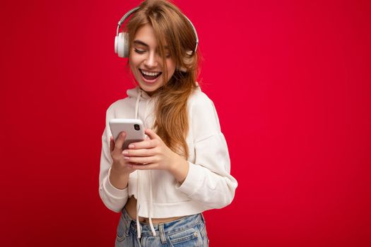 Photo of beautiful happy smiling young blonde woman wearing white hoodie isolated over colourful background wall with copy space for text wearing white wireless bluetooth earphones listening to cool music and holding mobile phone and texting sms.