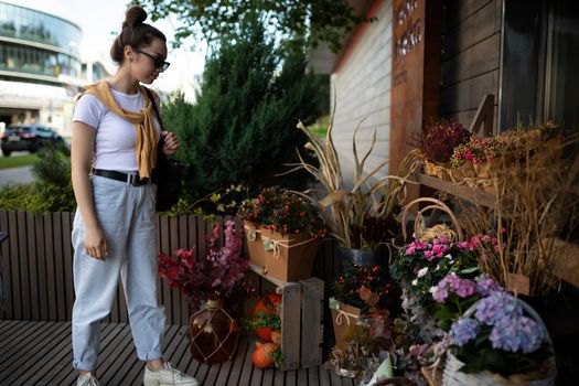 Photo of european beautiful attractive brunette woman wearing stylish white t-shirt and blue jeans chooses flowers as a gift in a street garden stall in the town.