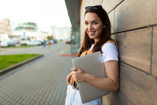 Photo of pretty attractive beautiful smiling happy positive charming young brunette young woman holding gray computer laptop wearing stylish clothes in white t-shirt and light blue jeans in the street in the town near brown wall.