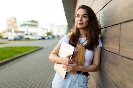 Photo of pretty attractive beautiful wonderful positive charming young brunette young woman holding grey computer laptop wearing stylish clothes in white t-shirt and light blue jeans in the street in the town near brown wall.