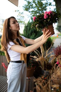 Vertical photo of european beautiful attractive positive happy brunette woman wearing stylish white t-shirt and blue jeans chooses flowers as a gift in a street garden stall in the town.