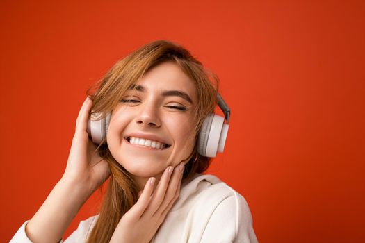 Close-up photo of beautiful happy smiling young blonde woman wearing white hoodie isolated over colourful background wall with copy space for text wearing white wireless bluetooth earphones listening to cool music and having fun.
