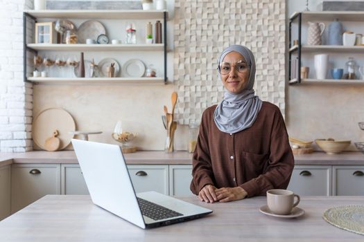 Portrait of young Muslim woman in hijab, woman in glasses looking at camera studying at home sitting in kitchen, female student using laptop for remote online learning.