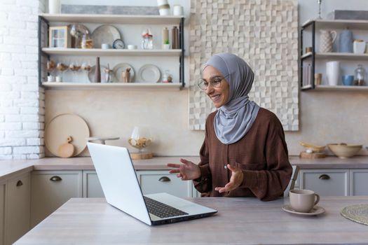 Young beautiful Muslim woman in hijab using laptop for video call, woman at home sitting in kitchen talking with friends remotely smiling at computer web camera