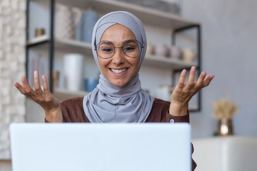 Video call online meeting with friends, young beautiful muslim woman in hijab looking at camera and smiling talking with friends remotely, woman at home in kitchen using laptop.