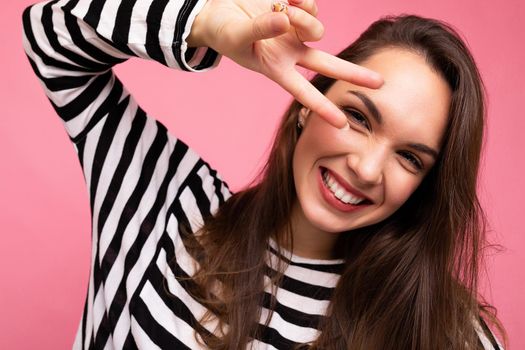 Close-up photo of young european positive smiling winsome happy beautiful brunette woman with sincere emotions wearing casual striped pullover and showing peace gesture eith two fingers isolated on pink background with copy space for text.