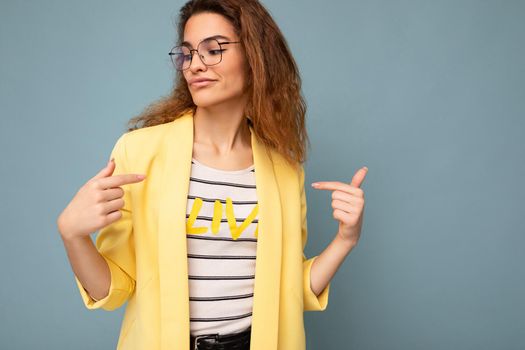 Photo of young positive sexy self-confident beautiful brunette curly woman with sincere emotions wearing casual yellow jacket and optical glasses isolated on blue background with copy space.