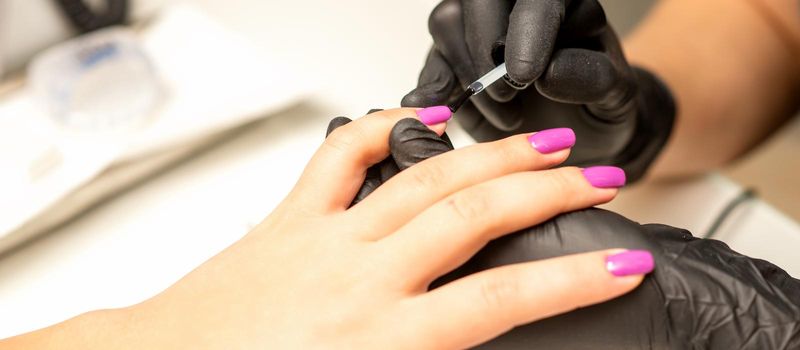 Professional manicure. A manicurist is painting the female nails of a client with purple nail polish in a beauty salon, close up. Beauty industry concept