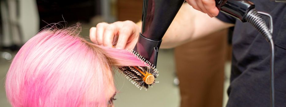 Drying short pink hair of young caucasian woman with a black hairdryer and black round brush by hands of a male hairdresser in a hair salon, close up