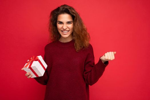 Photo of beautiful attractive charming positive happy funny young brunette curly woman wearing stylish dark red sweater isolated over red background wall with free space for text and holding white gift box with red ribbon.