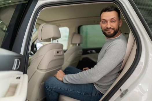 a customer in a car dealership sits in the back seat of a car with a smile on his face.