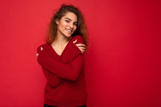 Photo of beautiful attractive charming positive happy smiling young brunette curly woman wearing stylish dark red sweater isolated over red background wall with free space for text.