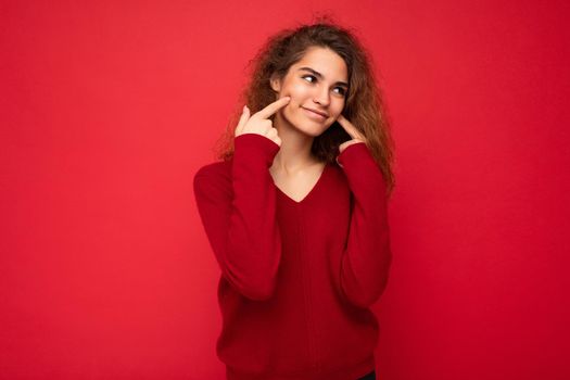 Photo of young positive smiling happy attractive brunette curly woman with sincere emotions wearing casual red sweater isolated on red background with copy space.