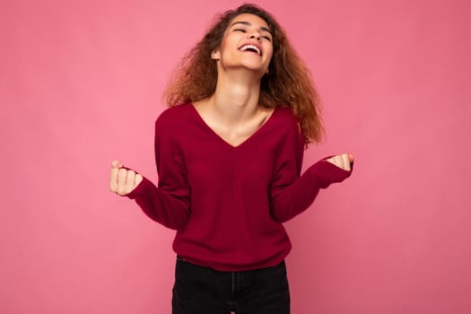 Photo of young emotional happy positive beautiful brunette curly woman with sincere emotions wearing casual pink pullover isolated over pink background with copy space and celebrating victory.