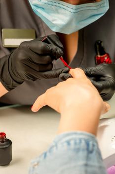 Manicure varnish painting. Close-up of a manicure master wearing rubber black gloves applying red varnish on a female fingernail in the beauty salon