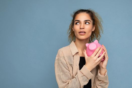 Photo of cute young beautiful attractive woman with dark curly hair in a ponytail wearing casual beige shirt isolated over blue background with copy space and holds pink piggy box for coins.