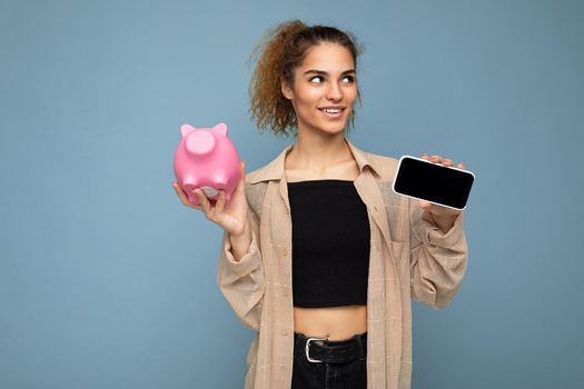 Portrait of happy positive smiling young beautiful attractive brunette curly woman with sincere emotions wearing stylish beige shirt isolated over blue background with copy space, holds pink pig money box and shows mobile phone with empty screen display. Payment concept.