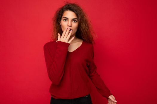Photo of youngshocked amazed attractive brunette curly woman with sincere emotions wearing casual red sweater isolated on red background with copy space.