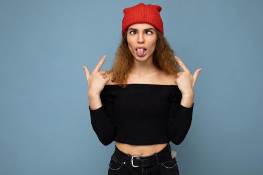 Photo of young positive beautiful brunette curly woman with sincere emotions wearing black crop top and red hat isolated on blue background with copy space and grimacing.