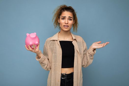 Portrait photo of sincere emotional dissatisfied and frustrated with something young beautiful woman with dark curly hair in a ponytail wearing casual beige shirt isolated over blue background with empty space and holds pink pig penny bank. Moneybox concept.